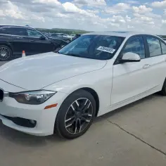 Biały sedan BMW na placu z innymi samochodami, w tym czarnym SUV-em. W tle widać więcej pojazdów, a niebo jest zachmurzone. To nie są amerykańskie samochody, ale europejskie klasyki.