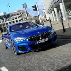 Błękitne BMW M850i sunie po miejskim asfalcie, mijając nowoczesne budynki.