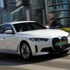 Białe BMW i4 E40 jedzie ulicą miasta, mijając nowoczesne budynki.