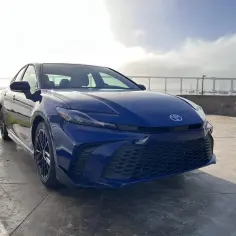 Błękitna Toyota Camry Hybrid zaparkowana nad wodą, z mostem w tle.