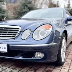 Niebieski Mercedes-Benz e211 z grillem i logo gwiazdy, zaparkowany na brukowanym podjeździe.