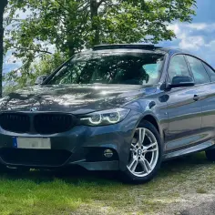 Ciemnoszary BMW F34 z czarnym grillem i felgami stoi na trawie pod drzewami.