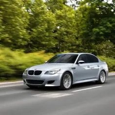 Srebrne BMW e60 m5 pędzi po drodze, otoczone rozmytą zielenią drzew.