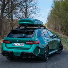 Zielone BMW M5 Touring z boxem dachowym gotowe na rodzinną podróż po leśnej drodze.