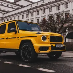 Żółty Mercedes G63 z czarnymi felgami na tle eleganckiego budynku.