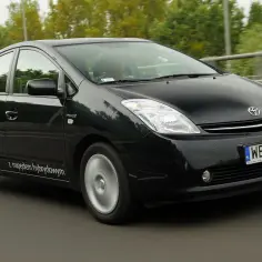 Czarny Toyota Prius 2 z napędem hybrydowym jedzie po drodze, otoczony zielenią.