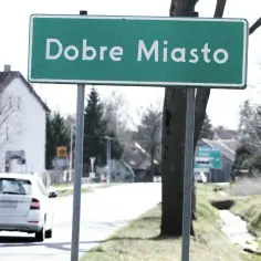 Zielony znak miejscowości "Dobre Miasto" wita podróżnych. W tle biały samochód i domy.