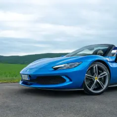 Niebieski kabriolet Ferrari 296 GTS na tle zielonych wzgórz i chmur.
