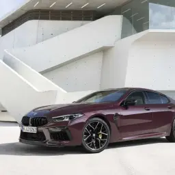 Eleganckie, ciemnoczerwone BMW M8 Gran Coupe prezentuje się na tle nowoczesnej architektury.