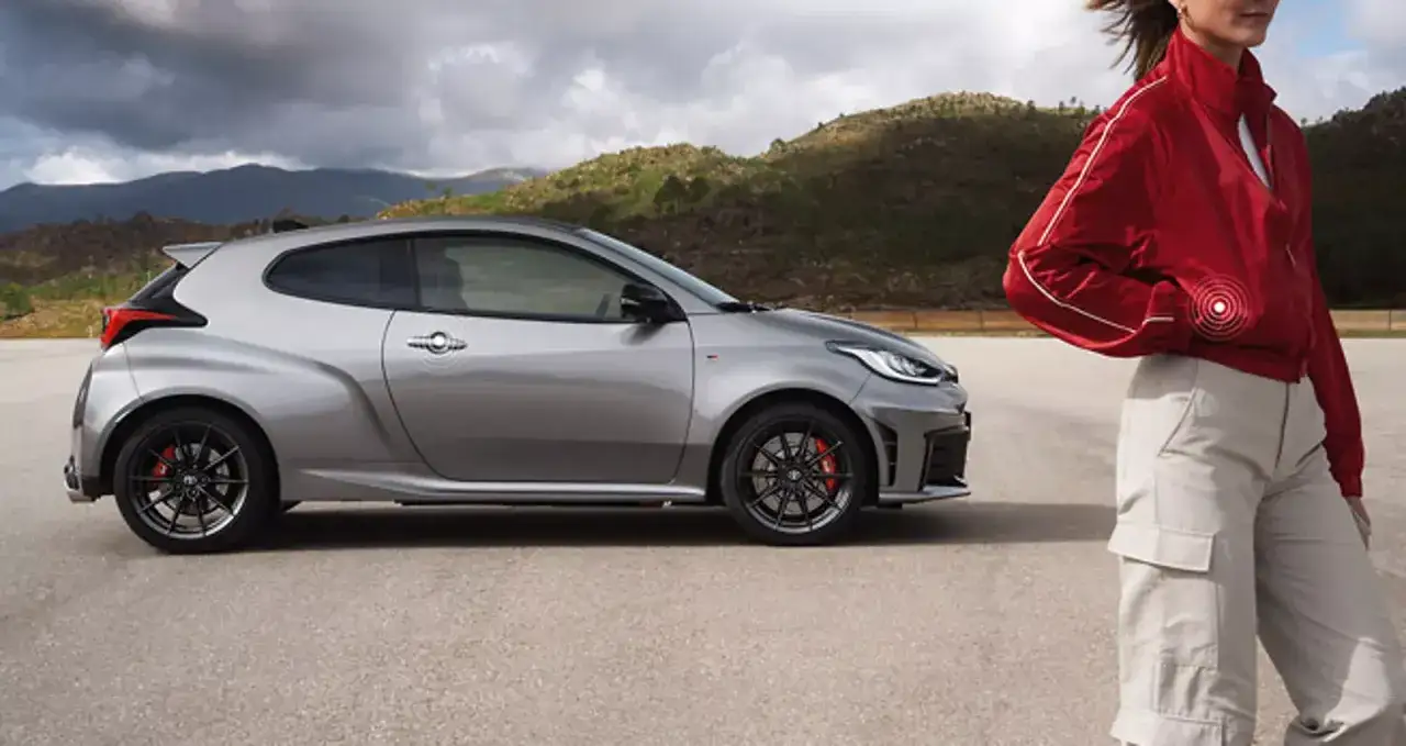 Srebrna toyota sportowa z czerwonymi zaciskami hamulcowymi na tle górskiego krajobrazu. Obok niej stoi kobieta w czerwonej kurtce.