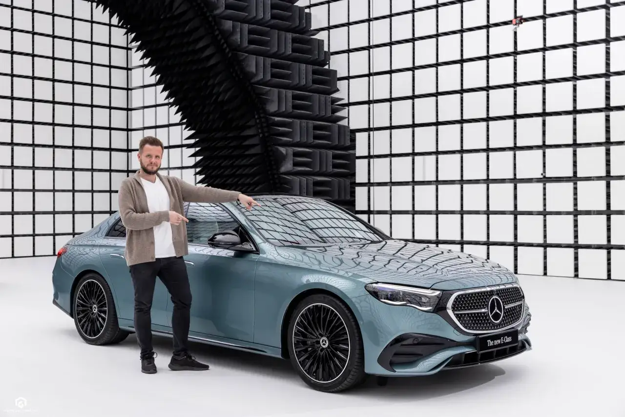 Mężczyzna prezentuje nowy, elegancki mercedes w214 w unikalnym kolorze. Tło tworzy nowoczesna ściana z białych kwadratów i czarnych paneli akustycznych.