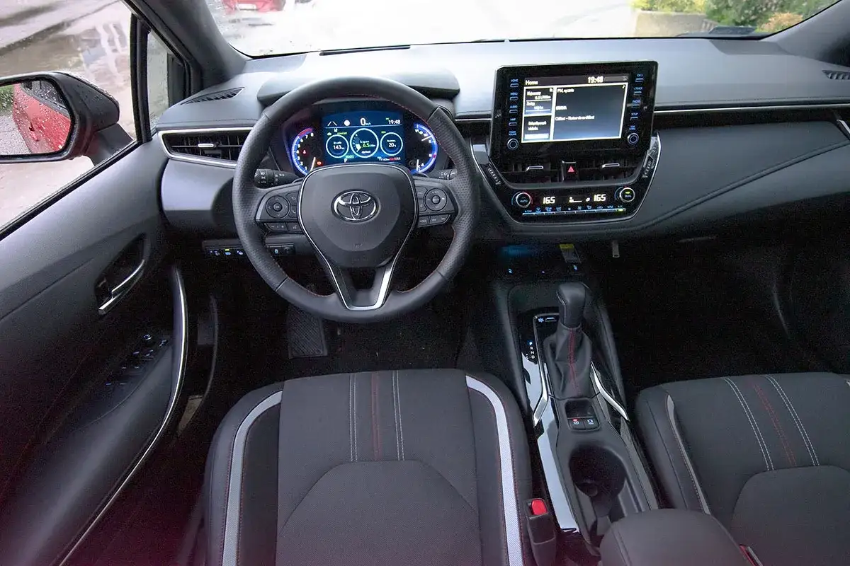 Wnętrze Toyota Corolla GR Sport: cyfrowe zegary, ekran multimediów, sportowa kierownica i fotele z czerwonymi przeszyciami.