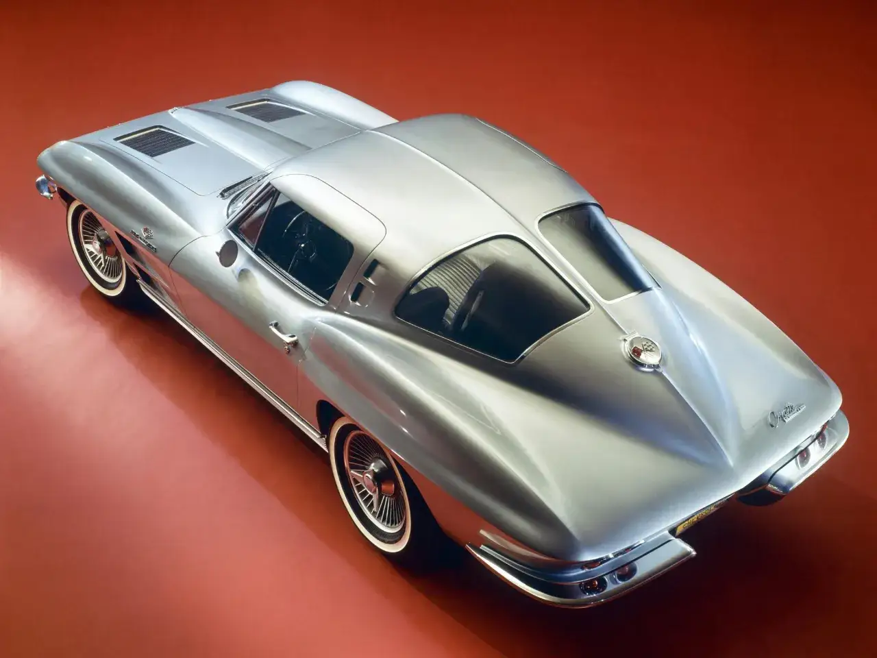 Srebrny corvette c2 Sting Ray z lat 60. na czerwonym tle, z widokiem z góry i lekko z boku.