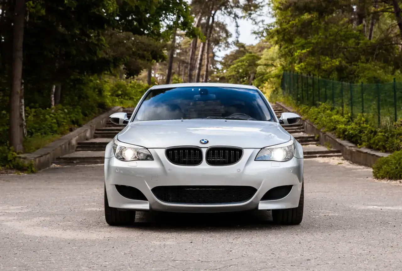 Srebrny BMW e60 m5 z włączonymi światłami, zaparkowany na drodze wśród drzew, z kamiennymi schodami w tle.