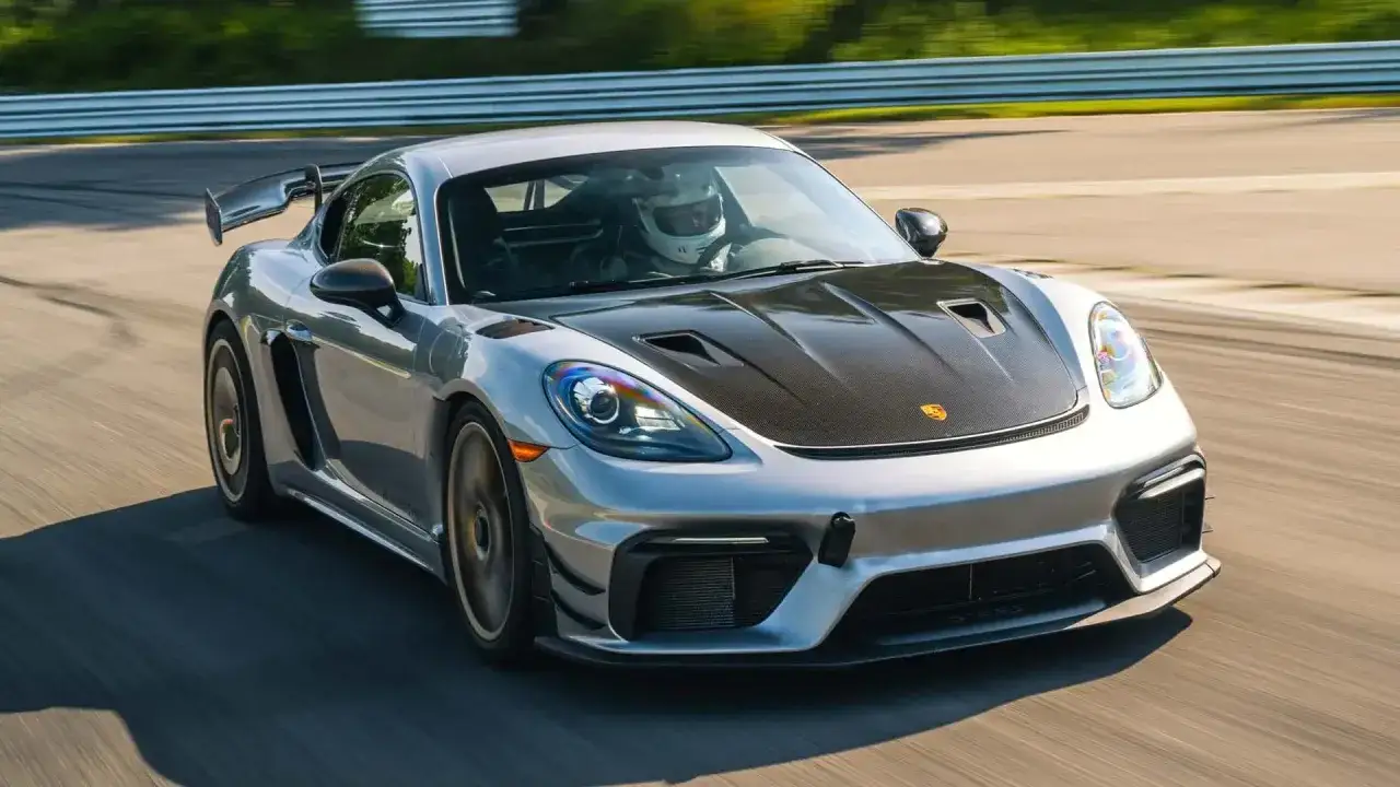 Srebrny Porsche GT4 RS z karbonową maską pędzi po torze wyścigowym, kierowca w kasku skupiony na drodze.