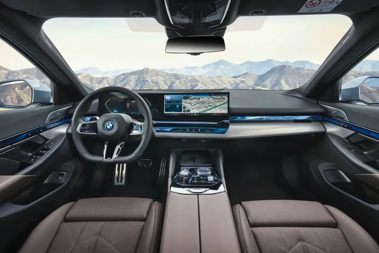 Wnętrze BMW i5 z panoramicznym ekranem, nowoczesną kierownicą i brązową skórzaną tapicerką. Widok na górzysty krajobraz przez przednią szybę.