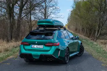 Zielone BMW M5 Touring z boxem dachowym gotowe na rodzinną podróż po leśnej drodze.